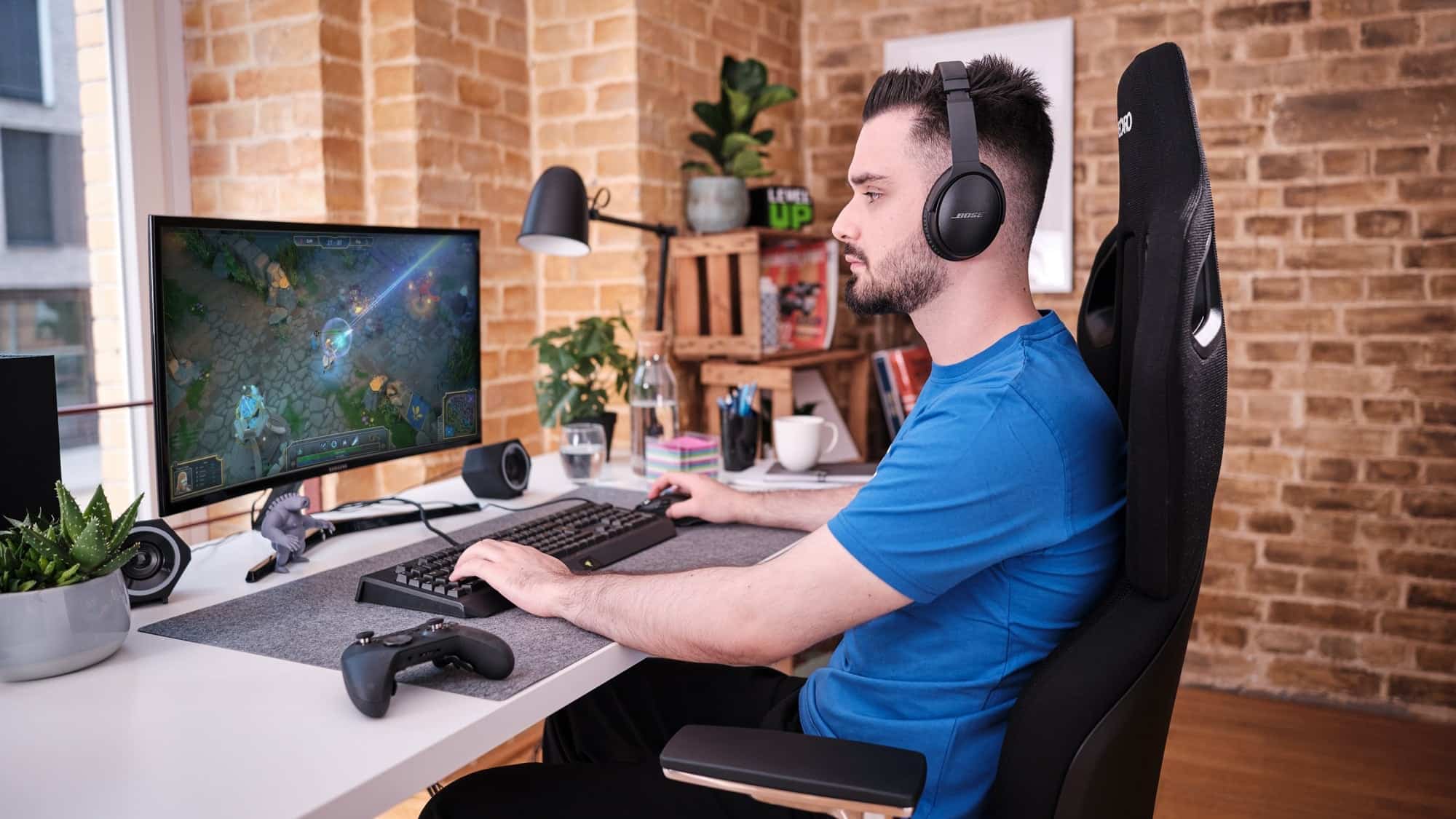 A man wearing headphones sits at a desk playing a multiplayer online game on a computer monitor in a modern home office.